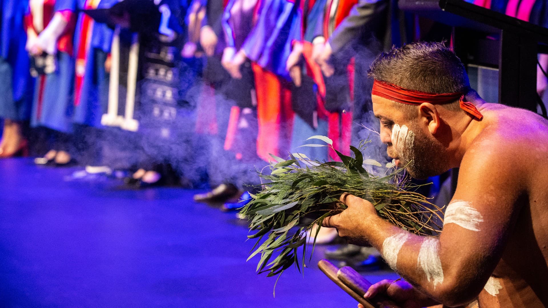 UOW’s new-look student graduations strengthen the connection with Country. Image of first nations person performing a smoking ceremony showing how the university is managing their Australia Day brand strategy.