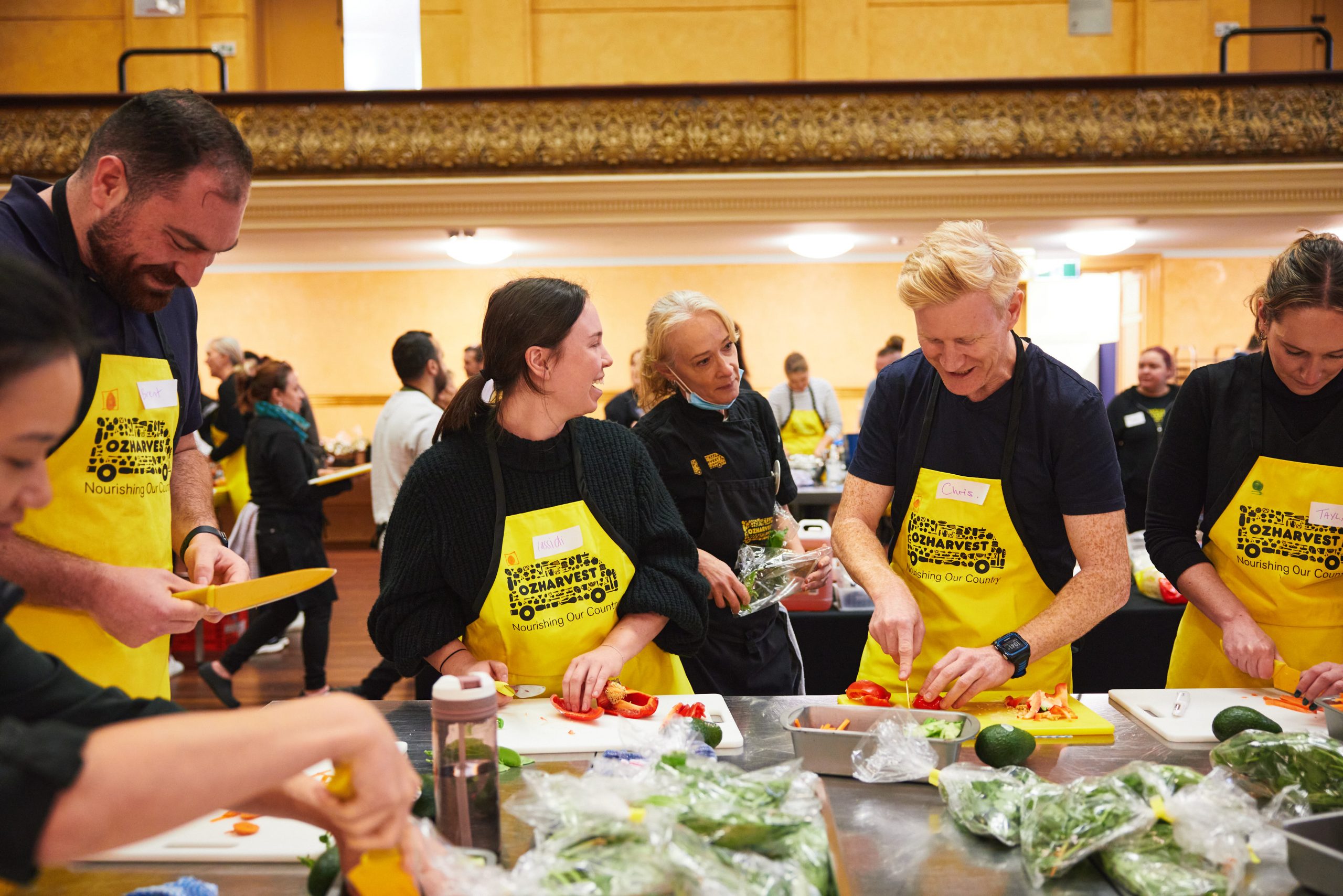 Several people preparing food and smiling, wearing Oz Harvest yellow aprons.